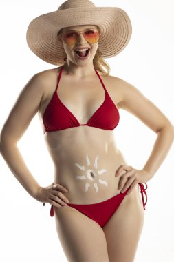 Kırmızı bikinili güzel, gülümseyen sarışın kadın. Beyaz stüdyo arka planında, göbeğinde güneş şeklinde kozmetik ürünler var..