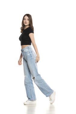 Yırtık kot pantolon ve siyah gömlek giyen genç bir manken beyaz stüdyo arka planında yürüyor. Yan, profilet görünümü