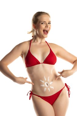 Kırmızı bikinili güzel, gülümseyen sarışın kadın. Beyaz stüdyo arka planında, göbeğinde güneş şeklinde kozmetik ürünler var..