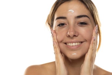 Genç, güzel, mutlu bir kadın beyaz bir stüdyoda yüzüne krem sürüyor.