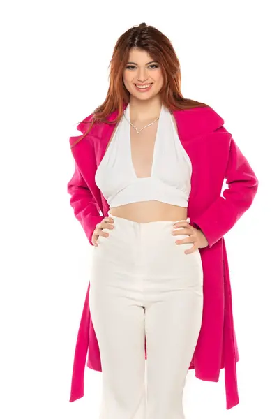 stock image Fashion style studio portrait of beautiful young woman. White wide leg pants and pink coat. Model sitting and posing against white studio background