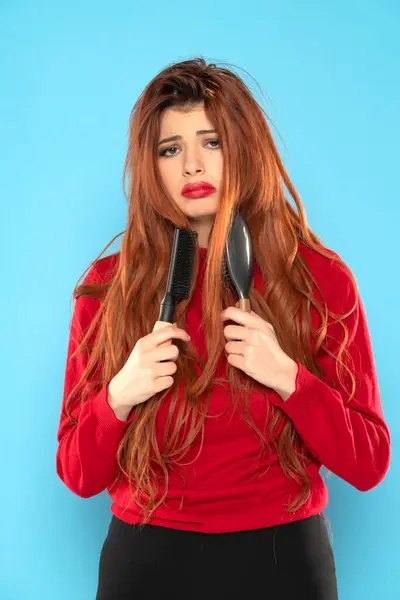 nervous young redhead woman with brush stuck in her messy hair on a blue studio background