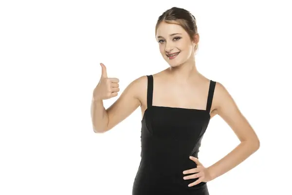 年轻的女人穿着黑色的连衣裙站在白色的工作室背景上 赞成用手做一个积极的手势 微笑着竖起大拇指 为成功感到高兴 赢的手势 免版税图库图片