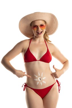 Kırmızı bikinili güzel, gülümseyen sarışın kadın. Beyaz stüdyo arka planında, göbeğinde güneş şeklinde kozmetik ürünler var..