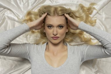 Moda portresi. Mavi gözlü, uzun sarı saçlı, beyaz saten üzerinde yatan genç bir model.