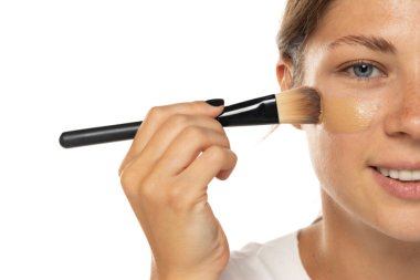 Beyaz bir stüdyo arka planına sıvı makyaj temeli uygulayan güzel bir genç kadının yarı portresi.