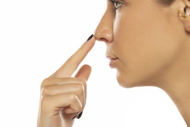 Genç ciddi bir kadın, beyaz stüdyo arka planında parmaklarıyla burnuna dokunuyor.