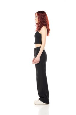 Stüdyoda siyah kısa bluz ve beyaz stüdyo arka planında siyah pantolon ile poz veren çekici genç bir kızıl kadın. Yan görünüm