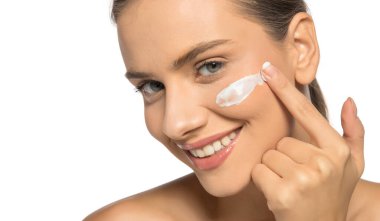 Güzel, genç, gülümseyen bir kadın beyaz bir stüdyo geçmişine yüz kremi sürüyor.