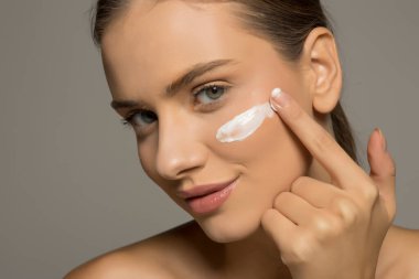 Güzel, genç, gülümseyen bir kadın gri bir stüdyo arka planına yüz kremi sürüyor.