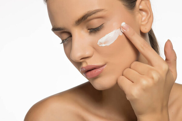 Красивая молодая женщина наносит крем для лица на белом фоне студии