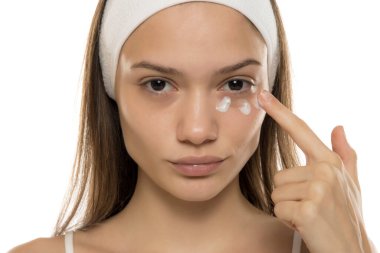 Güzellik. Genç bir kadın beyaz bir arka planda gözlerinin altına kozmetik krem sürüyor.