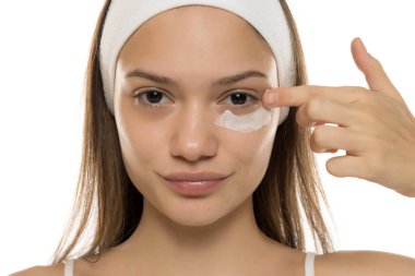 Güzellik. Genç, gülümseyen bir kadın beyaz bir arka planda gözlerinin altına makyaj kremi sürüyor.