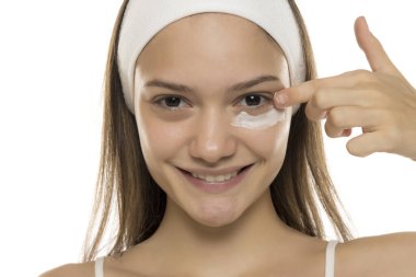 Güzellik. Genç, gülümseyen bir kadın beyaz bir arka planda gözlerinin altına makyaj kremi sürüyor.