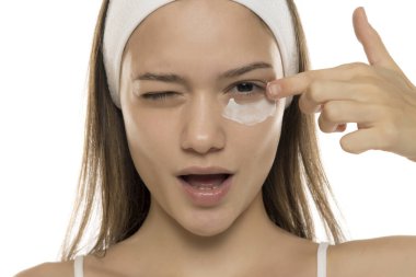 Güzellik. Genç, gülümseyen bir kadın beyaz bir arka planda gözlerinin altına makyaj kremi sürüyor.