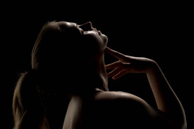 Sarışın at kuyruklu, başı arkaya eğilmiş, yüzüne dokunan bir kadının dramatik silueti. Düşük tuşlu ışıklandırma gölgeleri koyu arkaplana karşı.