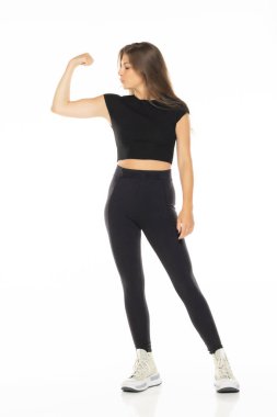 Siyah aktif giysili kadın, kaslarını esnetiyor, beyaz arka planda beyaz spor ayakkabılı güç ve güven gösteriyor..