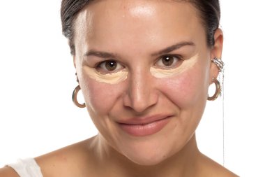 Beyaz stüdyo arka planında gözlerinin altında kapatıcı ile poz veren gülümseyen kadın.