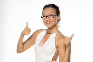 Beyaz elbiseli, yakalı, gözlüklü, neşeli Asyalı kadın. İki elinle baş parmağını kaldırman, geniş gülümsemen. Beyaz arkaplan karşısında stüdyo görüntüsü.