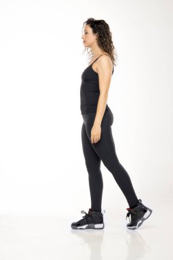 Siyah atlet tişörtlü, spor ayakkabılı, kıvırcık saçlı genç bir kadın profili. Beyaz arka plan karşısında doğal duruş sergiliyor..