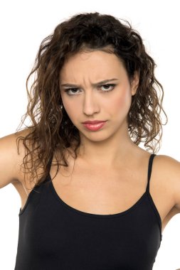 Siyah atlet giyen kıvırcık saçlı, beyaz arka plana karşı somurtkan ve şüpheci ifadeler gösteren genç bir kadının yakın plan portresi..