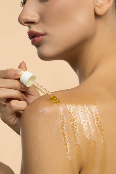 Крупный план женщины, наносящей оливковое масло на плечо капельницей. На коже видны полосы масла, демонстрирующие естественное лечение кожи. Нейтральный студийный фон.