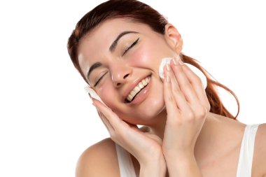 Güzel kızıl saçlı, gülümseyen kadın beyaz stüdyo arka planında pamukla yüzünü temizliyor. 