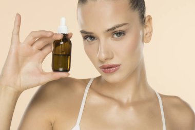 Yüzünün yanında kehribar rengi cam damlatıcı şişesi tutan güzel genç bir kadın bej arka planda cilt ürünü sergiliyor.