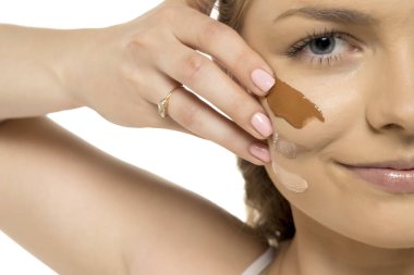 Yanağında üç temel gölgesi olan bir kadının yüzüne aşırı yakın çekim, beyaz arka planda makyaj rengi testini gösteriyor..