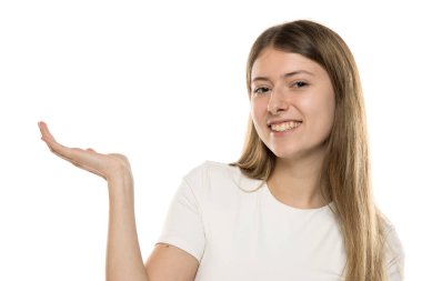 Uzun saçlı genç bir kadın gülümsüyor ve elini dışarı doğru uzatıyor. Parlak, minimalist bir ortamda dostane ve cana yakın bir tavır sergiliyor..