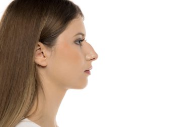 Bu görüntü, uzun kahverengi saçlı, narin yüz hatlarını ve temiz beyaz bir arkaplan karşısında sakin ifadesini sergileyen genç bir kadının yan profilini içeriyor..