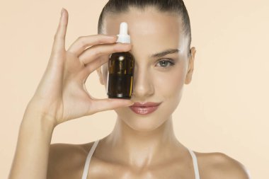 Yüzünün yanında kehribar rengi cam damlatan şişeyle gülümseyen güzel bir kadın bej arka planda cilt ürünü sergiliyor.
