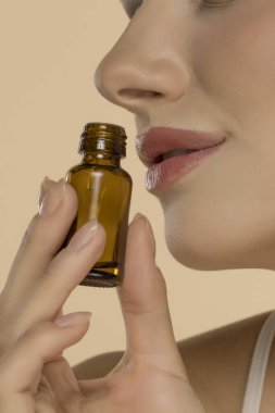 Gözleri kapalı bir kadın kehribar şişeyi burnuna dayayıp koku soluyor. Aromaterapi uygulamasını gösteren yakın profil görüntüsü. Tarafsız arkaplan, doğal makyaj.