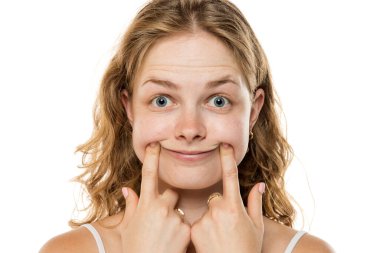 Dalgalı sarı saçlı genç kadın, beyaz arka plana karşı sahte duygular gösteren parmaklarıyla yanaklarını iterek gülümsemeye çalışıyor..