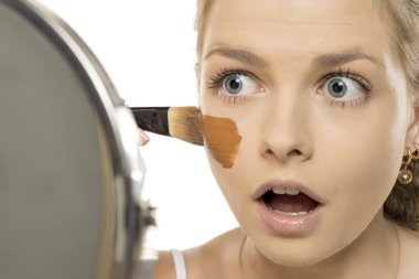 Cilt rengine uymayan temeli uygularken aynaya bakan şok olmuş genç bir kadın makyaj renk uyumsuzluğunu gösteriyor..