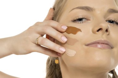 Yanağında üç farklı fondöten gölgesi olan tefekküre dalmış genç bir kadının yakın çekimi. Makyaj renkleriyle beyaz arkaplanı karşılaştırıyor..