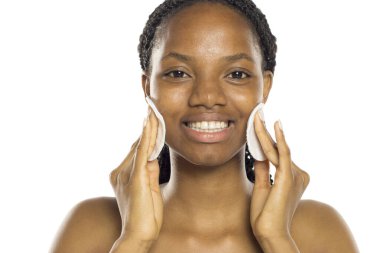 Siyah kadın yüzünü temizlemek için iki pamuk pedi kullanıyor. Beyaz arka planda bir stüdyoda gülümsüyor..