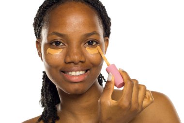 Gülümseyen siyah kadın göz altına koruyucu sürüyor, makyaja hazırlanıyor, beyaz arka planda stüdyo çekimi var..