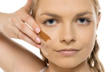 Yüzünde farklı fondöten örnekleri olan sakin yüzlü genç bir kadın beyaz arkaplana karşı elle makyaj gölgesi testi yapıyor..