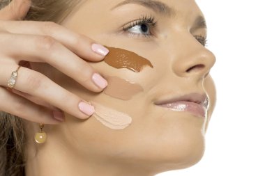 Yanağında üç farklı fondöten gölgesi olan tefekküre dalmış genç bir kadının yakın çekimi. Makyaj renkleriyle beyaz arkaplanı karşılaştırıyor..