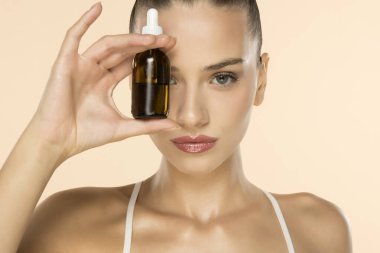 Yüzünün yanında kehribar rengi cam damlatıcı şişesi tutan güzel genç bir kadın bej arka planda cilt ürünü sergiliyor.