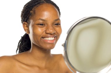 Siyah kadın aynadaki yansımasına gülümsüyor, teniyle mutlu, beyaz arka planda stüdyo çekimi var.