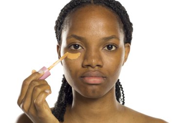 Siyah bir kadın göz altına sıvı kapatıcı sürüyor, temiz bir yüz makyajı için hazırlanıyor, beyaz arka planda stüdyo çekimi..