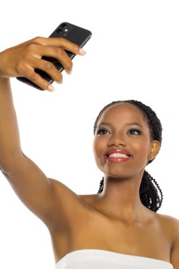 Makyajlı gülümseyen siyah kadın özçekim yapıyor, beyaz arka planda stüdyo çekimi var..