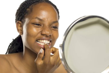 Siyah kadın aynada dişlerini kontrol ediyor. Parmak izi, gülümseyen yüz ifadesi, beyaz arka planda stüdyo çekimi.