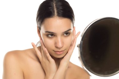 Esmer kadın yüzüne dokunuyor ve aynaya bakıyor. Stüdyo resmi beyaz arka planda.