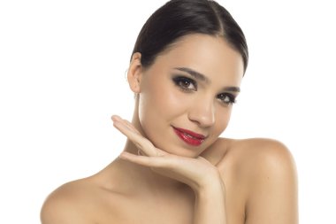 Beyaz arka planda, başı eğik ve eli çenesinin altında gülümseyen çekici bir kadının stüdyo çekimi.