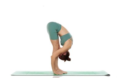Yeşil elbiseli genç bir kadının beyaz arka planda öne eğilmiş yoga pozu (uttanasana) verirken çekilmiş bir fotoğraf.