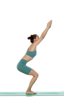 Yeşil elbiseli genç bir kadının beyaz arka planda yoga pozu (utkatasana) verirken çekilmiş bir fotoğraf.