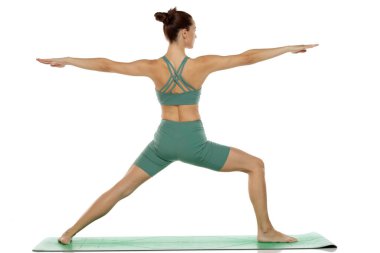Yeşil elbiseli genç bir kadının beyaz arka planda Savaşçı II yoga pozu (virabhadrasana II) verirken ki görüntüleri.
